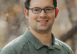 Smiling man with short dark hair and glasses wearing a green polo shirt outdoors.