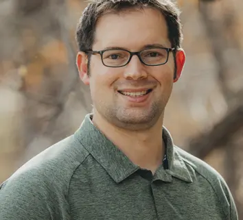 Smiling man with short dark hair and glasses wearing a green polo shirt outdoors.