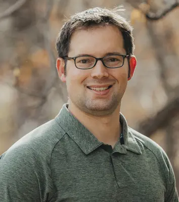 Smiling man with short dark hair and glasses wearing a green polo shirt outdoors.
