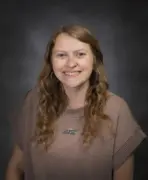 Portrait of a smiling woman with long, wavy light brown hair wearing a brown knit sweater against a dark studio backdrop, looking at the camera.