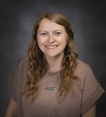 Portrait of a smiling woman with long, wavy light brown hair wearing a brown knit sweater against a dark studio backdrop, looking at the camera.