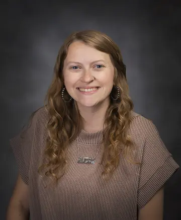 Portrait of a smiling woman with long, wavy light brown hair wearing a brown knit sweater against a dark studio backdrop, looking at the camera.