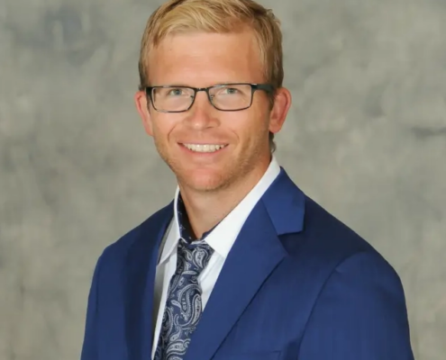 Smiling man in a blue suit and patterned tie, wearing glasses, posing for a studio portrait.