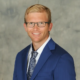 Smiling man in a blue suit and patterned tie, wearing glasses, posing for a studio portrait.
