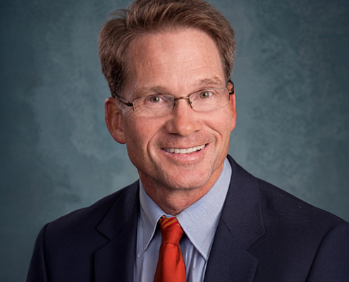 Professional headshot: man in a navy suit and red tie, wearing glasses, smiling against a blue backdrop.