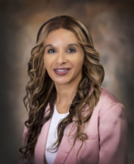 Professional headshot of a woman with long wavy light-brown hair, wearing a pink blazer and white top, smiling against a neutral studio background.