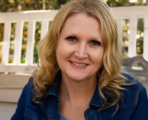 Smiling woman with wavy blonde hair wearing a denim jacket seated on a porch outdoors.