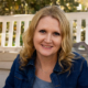 Smiling woman with wavy blonde hair wearing a denim jacket seated on a porch outdoors.