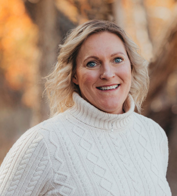 Portrait of a smiling woman with blonde hair wearing a white cable-knit turtleneck sweater, outdoors with autumn trees in the background.