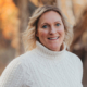 Portrait of a smiling woman with blonde hair wearing a white cable-knit turtleneck sweater, outdoors with autumn trees in the background.