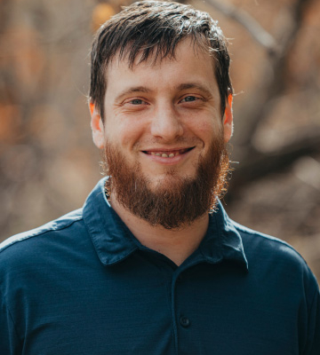 Smiling man with short brown hair and a beard, wearing a blue polo shirt, outdoors with blurred trees in the background.