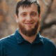 Smiling man with short brown hair and a beard, wearing a blue polo shirt, outdoors with blurred trees in the background.