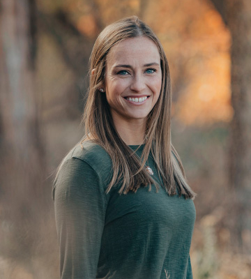 Portrait of a smiling woman with long brown hair in a dark green sweater, standing outdoors in a fall forest with warm sunlight