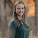 Portrait of a smiling woman with long brown hair in a dark green sweater, standing outdoors in a fall forest with warm sunlight
