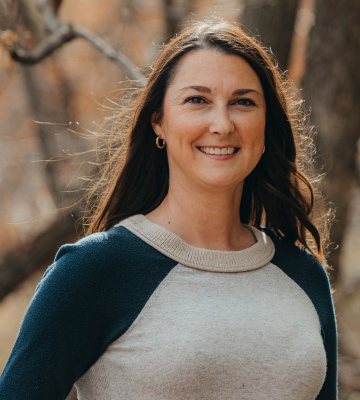 Smiling woman with long brown hair wearing a gray and navy sweater outdoors in a forest setting.