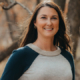 Smiling woman with long brown hair wearing a gray and navy sweater outdoors in a forest setting.