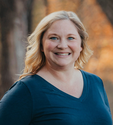 Portrait of a smiling woman with blonde hair wearing a blue V-neck shirt, outdoors with autumn trees in the background.