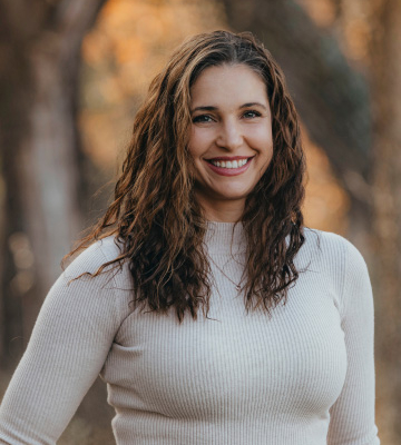 Smiling woman with wavy brown hair wearing a light sweater outdoors in autumn leaves behind her.
