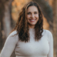 Smiling woman with wavy brown hair wearing a light sweater outdoors in autumn leaves behind her.