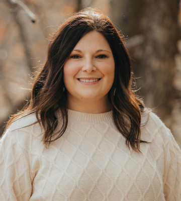Portrait of a smiling woman with dark hair wearing a cream knit sweater, outdoors with trees in the background.