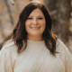 Portrait of a smiling woman with dark hair wearing a cream knit sweater, outdoors with trees in the background.