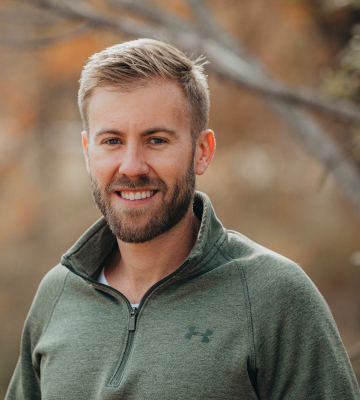 Portrait of a smiling man with blond hair and beard wearing a green pullover outdoors with autumn-colored trees behind him