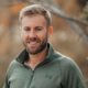 Portrait of a smiling man with blond hair and beard wearing a green pullover outdoors with autumn-colored trees behind him