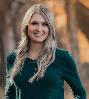 Smiling woman with long blonde hair wearing a teal top, standing outdoors with autumn foliage in the background.
