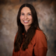 Portrait of a smiling woman with long dark hair wearing an orange blouse against a neutral studio backdrop.