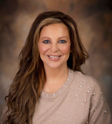 Studio portrait of a smiling woman with long wavy brown hair wearing a beige sweater with pearl embellishments, warm brown background.