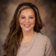 Studio portrait of a smiling woman with long wavy brown hair wearing a beige sweater with pearl embellishments, warm brown background.