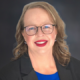 Professional headshot of a smiling woman with glasses, wearing a blue top and black blazer against a dark gradient background.