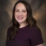 Portrait of a smiling woman with dark brown hair wearing a purple top, against a neutral studio backdrop.