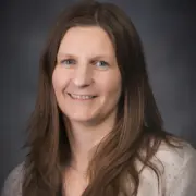 Portrait of a smiling woman with long brown hair, wearing a light sweater, against a dark studio backdrop.
