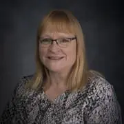 Smiling woman with blonde hair and glasses, wearing a patterned blouse, in a studio portrait against a dark background.