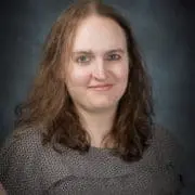 Portrait of a woman with shoulder-length brown hair wearing a patterned top, against a dark studio background.