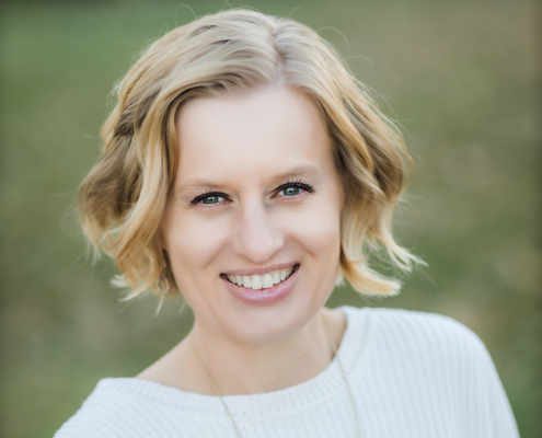 Smiling woman with short blonde hair outdoors, wearing a white sweater, looking at the camera.