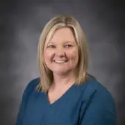 Smiling blonde woman in teal scrubs against a dark studio backdrop, portrait of a healthcare professional.