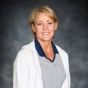 Smiling portrait of a woman with short blonde hair, wearing a white jacket over a striped shirt, against a dark studio background.