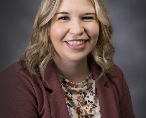 Smiling woman with blonde hair wearing a burgundy blazer and floral blouse, portrait against a gray backdrop.