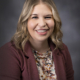 Smiling woman with blonde hair wearing a burgundy blazer and floral blouse, portrait against a gray backdrop.