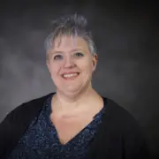 Smiling woman with short gray hair wearing a blue blouse and black cardigan, against a dark studio backdrop.