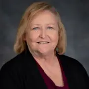 Portrait of a smiling older woman with blonde hair, wearing a black blazer and maroon blouse.