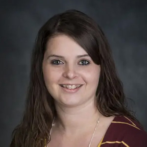 Portrait of a smiling woman with long brown hair, wearing a burgundy top, against a dark studio backdrop.