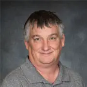 Portrait of a smiling middle-aged man with short gray hair wearing a gray polo shirt, against a neutral backdrop.