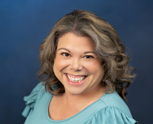 Portrait of a smiling woman with shoulder-length wavy gray-brown hair wearing a teal blouse against a blue background; professional headshot intent.