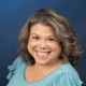 Portrait of a smiling woman with shoulder-length wavy gray-brown hair wearing a teal blouse against a blue background; professional headshot intent.