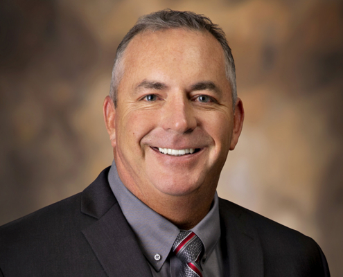 Smiling man in a dark suit and gray shirt, wearing a striped tie, against a blurred brown backdrop.