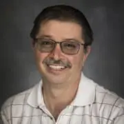 Portrait of a smiling man with glasses and a mustache, wearing a white checkered polo shirt, against a dark studio backdrop.