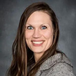 Portrait of a smiling woman with long brown hair wearing a gray sweater, against a dark gray backdrop.